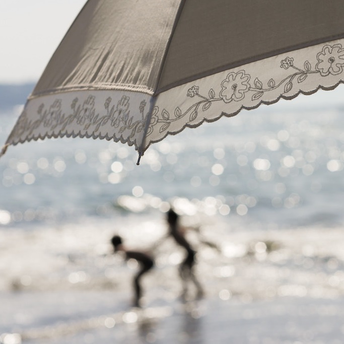 A photograph taken at a beach with the following elements and composition: In the foreground, a close-up view of a light-colored beach umbrella with intricate floral embroidery on the edge. The umbrella is positioned towards the top and right side of the image, partially covering the upper part of the scene. In the background, slightly out of focus, two children are playing in the shallow waves of the ocean. The ocean water is shimmering with sunlight reflections, creating a bokeh effect. The children are in the middle left part of the image, with one child standing and the other bending down. The overall mood is serene and playful, with a soft, dreamy quality to the photograph.