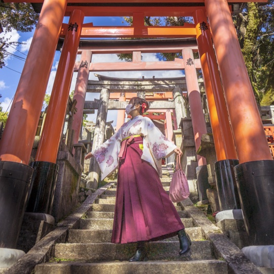 A woman in traditional Japanese attire, standing on stone steps leading to a series of Torii gates at Fushimi Inari Shrine in Kyoto. She is wearing a white kimono with floral patterns and a maroon hakama, holding a small bag in one hand. The sunlight filters through the trees, casting shadows and highlighting the vibrant red and orange of the Torii gates. The composition emphasizes the woman's elegance and the spiritual atmosphere of the shrine, creating an impression of grace, tradition, and serenity.